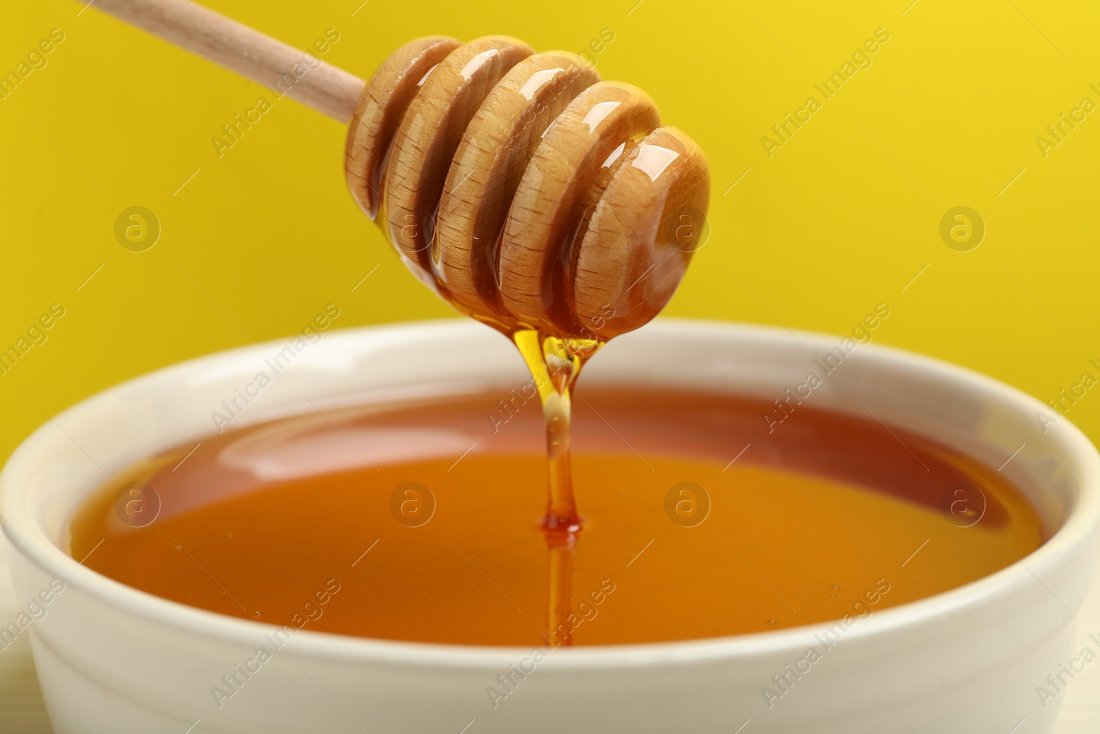 Photo of Pouring tasty honey from dipper into bowl against yellow background, closeup