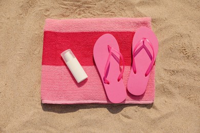 Beach towel with slippers and sunscreen on sand, top view