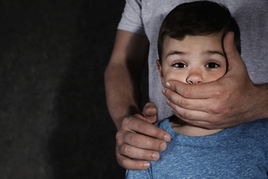 Adult man covering scared little boy's mouth on dark background, space for text. Child in danger