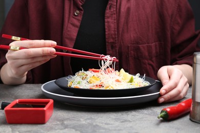 Photo of Stir-fry. Woman with chopsticks eating tasty rice noodles with meat and vegetables at grey textured table, closeup