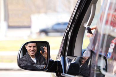 Photo of Man with key looking into side view mirror of modern car outdoors. Buying new auto