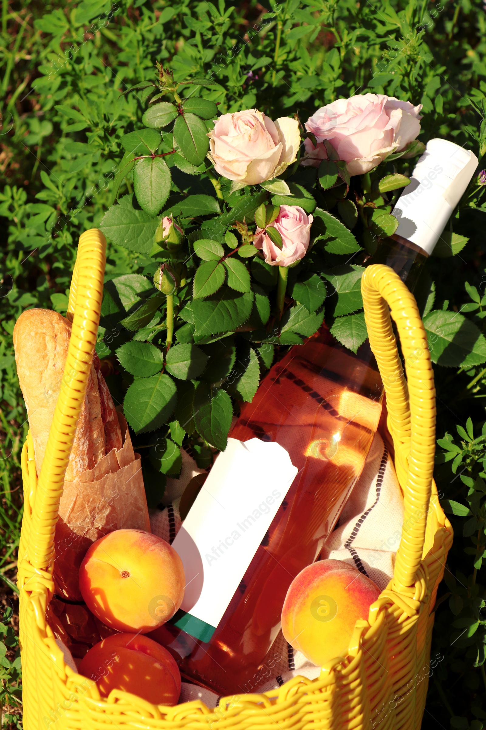 Photo of Yellow wicker bag with roses, peaches, baguette and wine outdoors, closeup