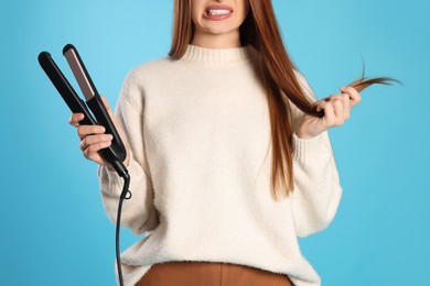 Photo of Upset young woman with flattening iron on light blue background, closeup. Hair damage