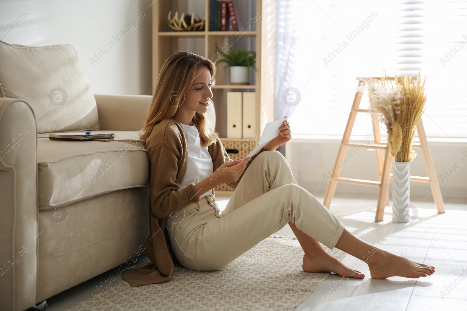 Photo of Happy woman reading letter while sitting on floor near sofa at home