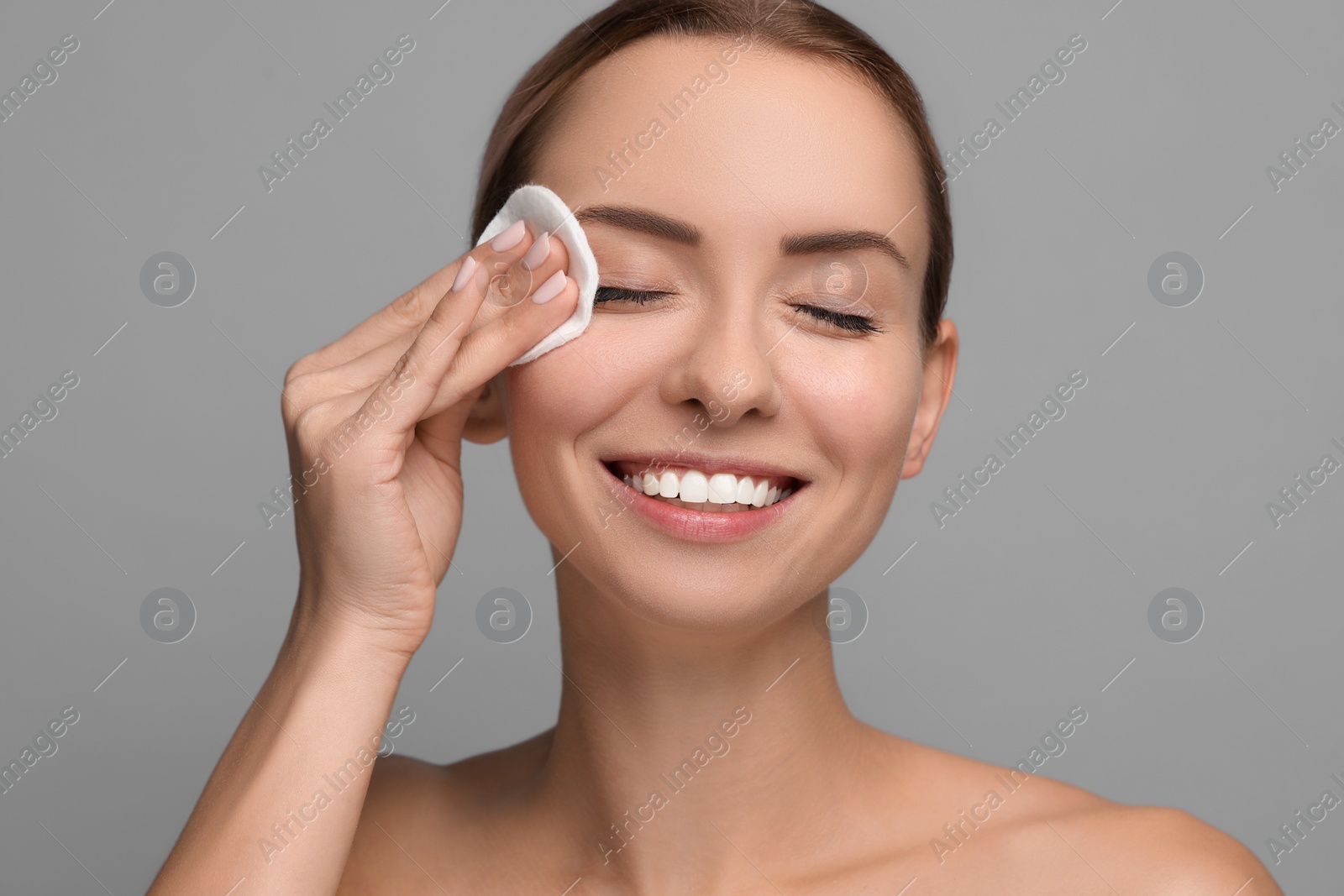 Photo of Smiling woman removing makeup with cotton pad on grey background, closeup