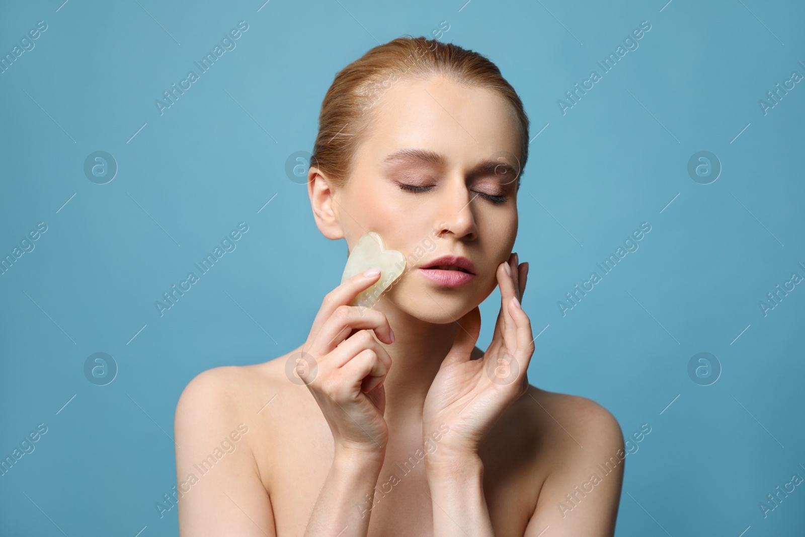 Photo of Beautiful young woman doing facial massage with gua sha tool on blue background