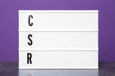 Photo of Light box with letters CSR on table against purple background. Corporate social responsibility