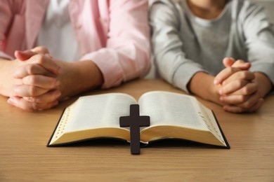 Boy praying together with his godparent at wooden table, focus on Bible and cross