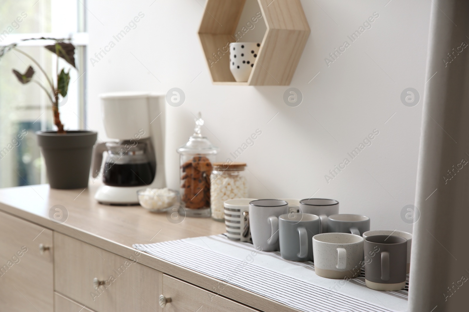 Photo of Modern coffeemaker and cups on commode near light wall indoors