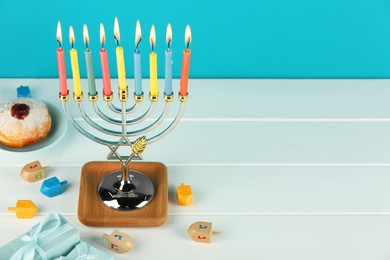 Photo of Hanukkah celebration. Menorah with burning candles, dreidels, donut and gift boxes on white wooden table against light blue background. Space for text