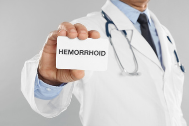 Doctor holding business card with word HEMORRHOID on light grey background, closeup