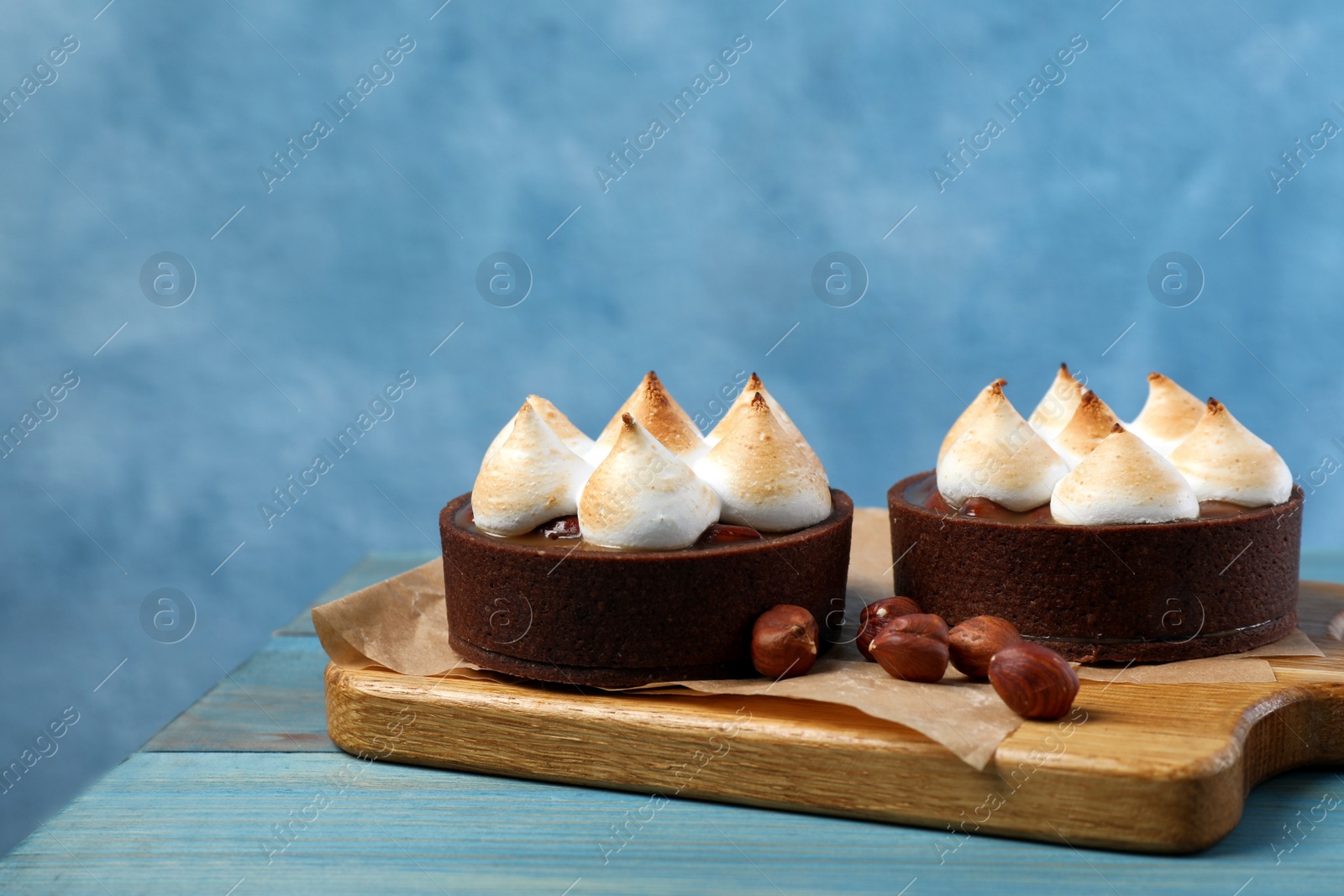 Photo of Delicious salted caramel chocolate tarts with meringue and hazelnuts on light blue wooden table, space for text