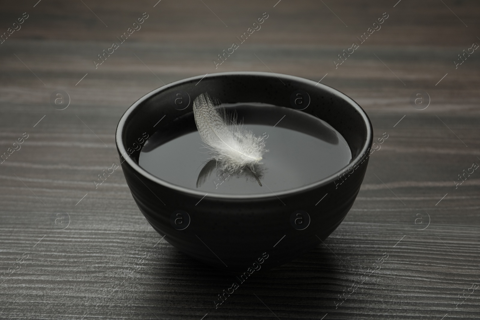 Photo of Black bowl with water and white feather on dark wooden table