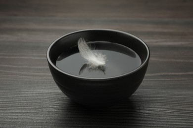 Photo of Black bowl with water and white feather on dark wooden table