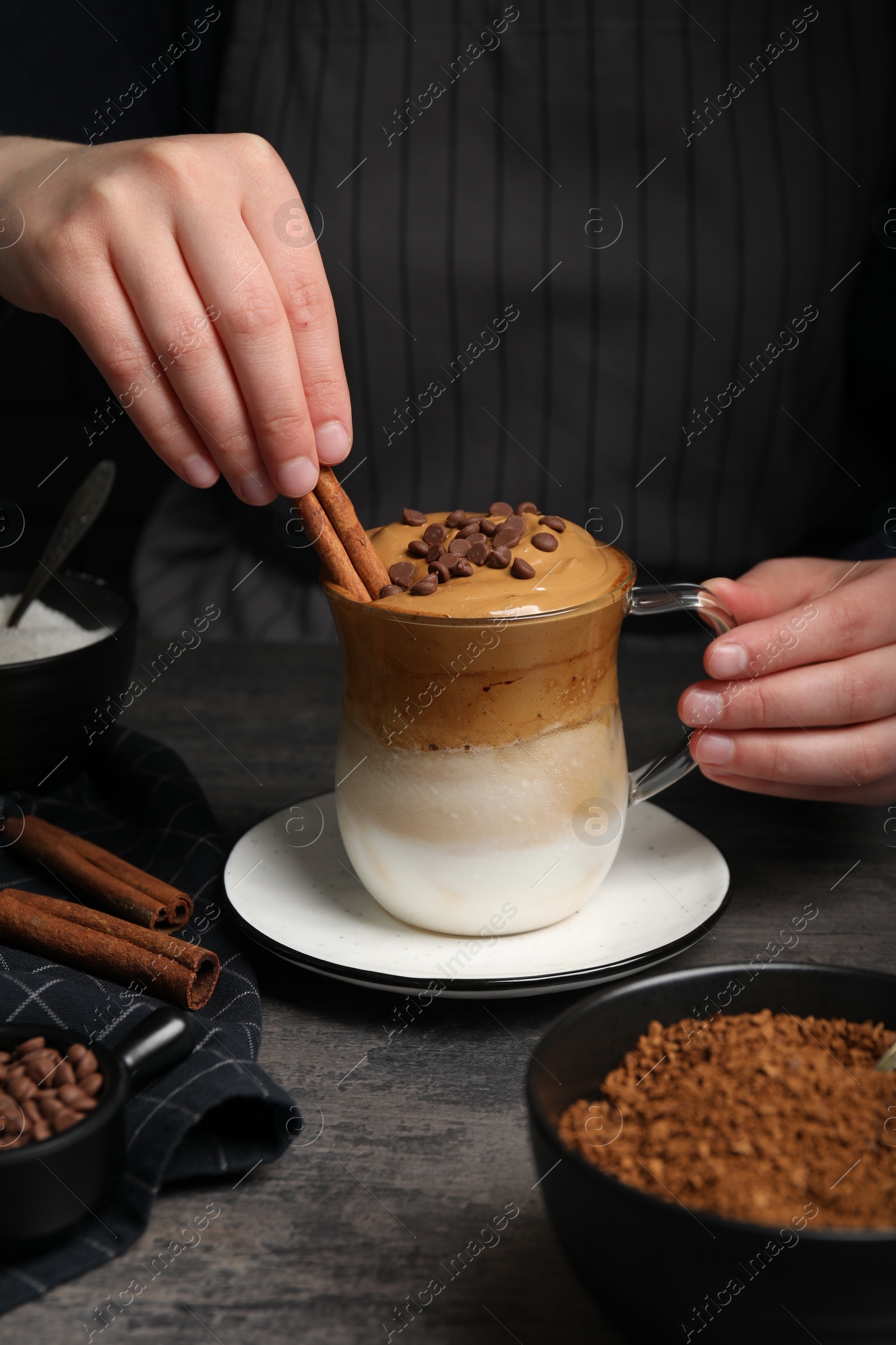 Photo of Woman decorating dalgona coffee with cinnamon sticks at grey table, closeup