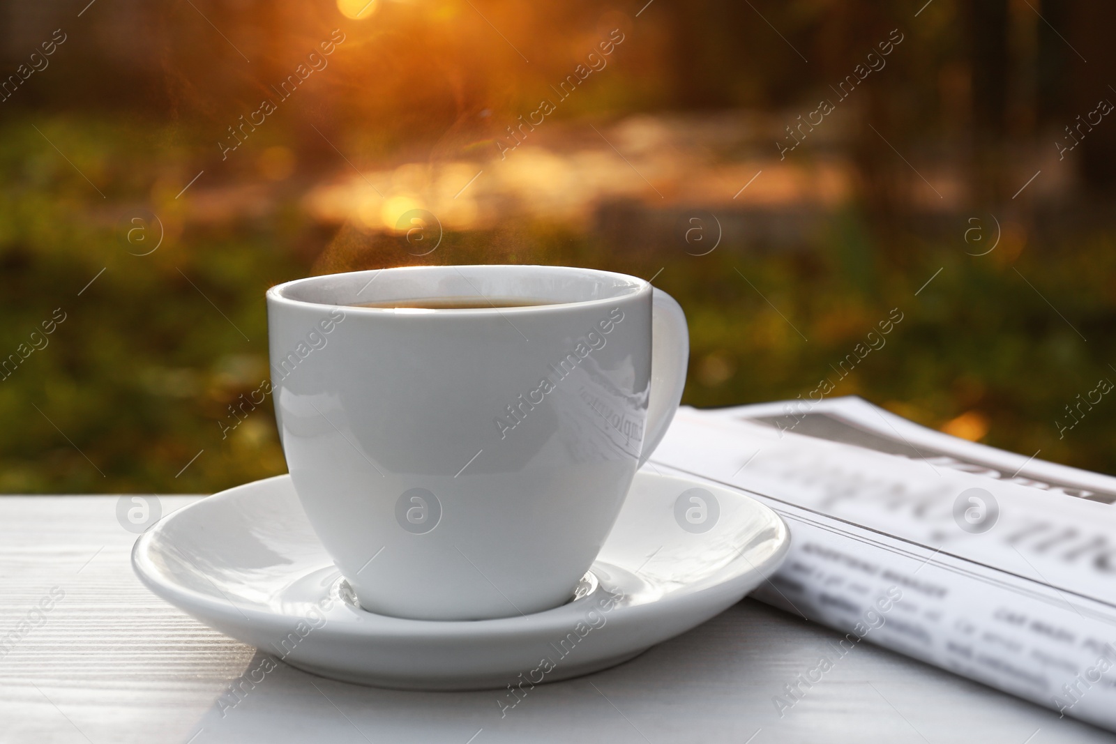 Photo of White cup with coffee and newspaper on wooden table in morning outdoors, closeup