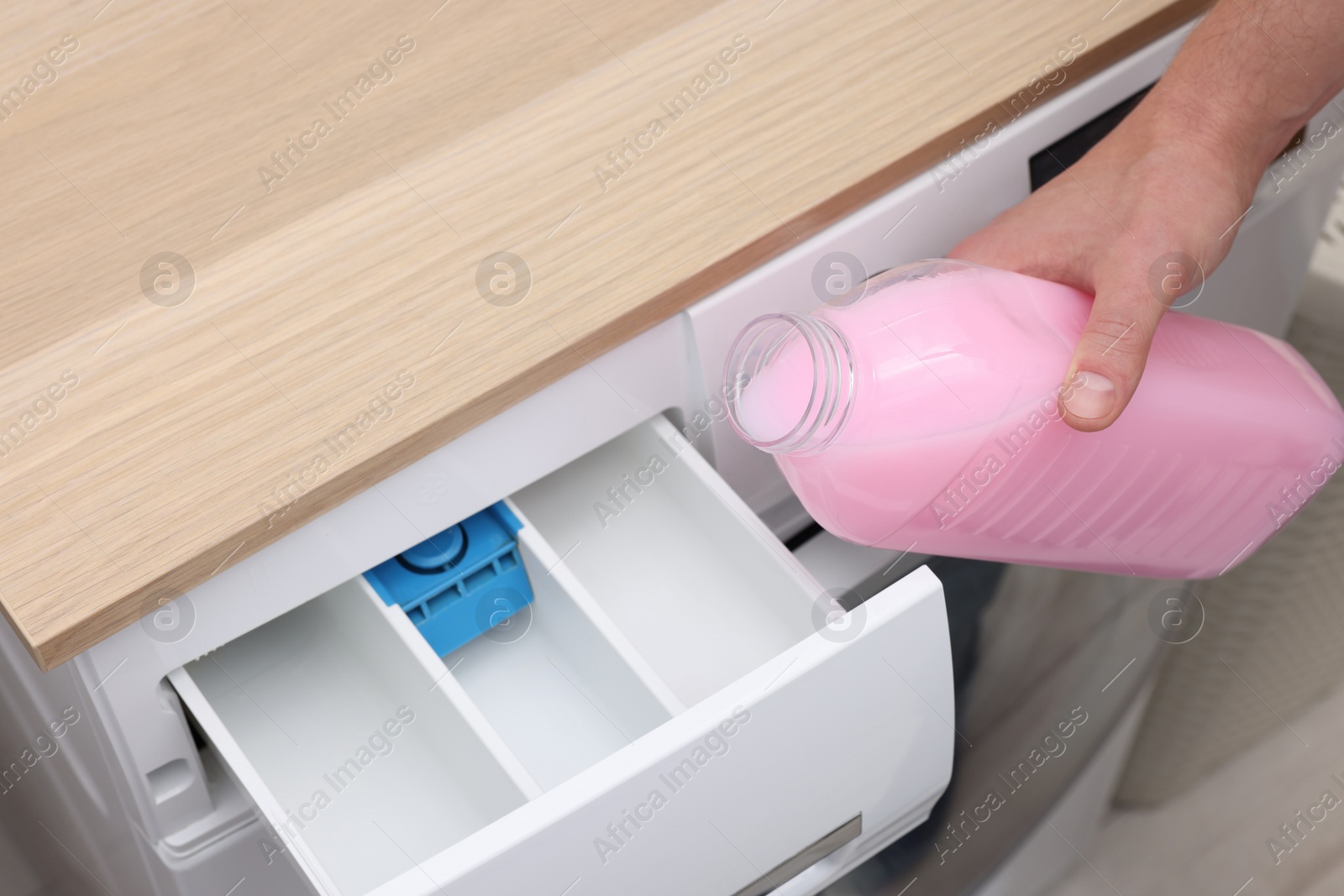 Photo of Man pouring fabric softener from bottle into washing machine indoors, closeup