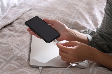 Woman connecting charger cable to smartphone near laptop on bed, closeup