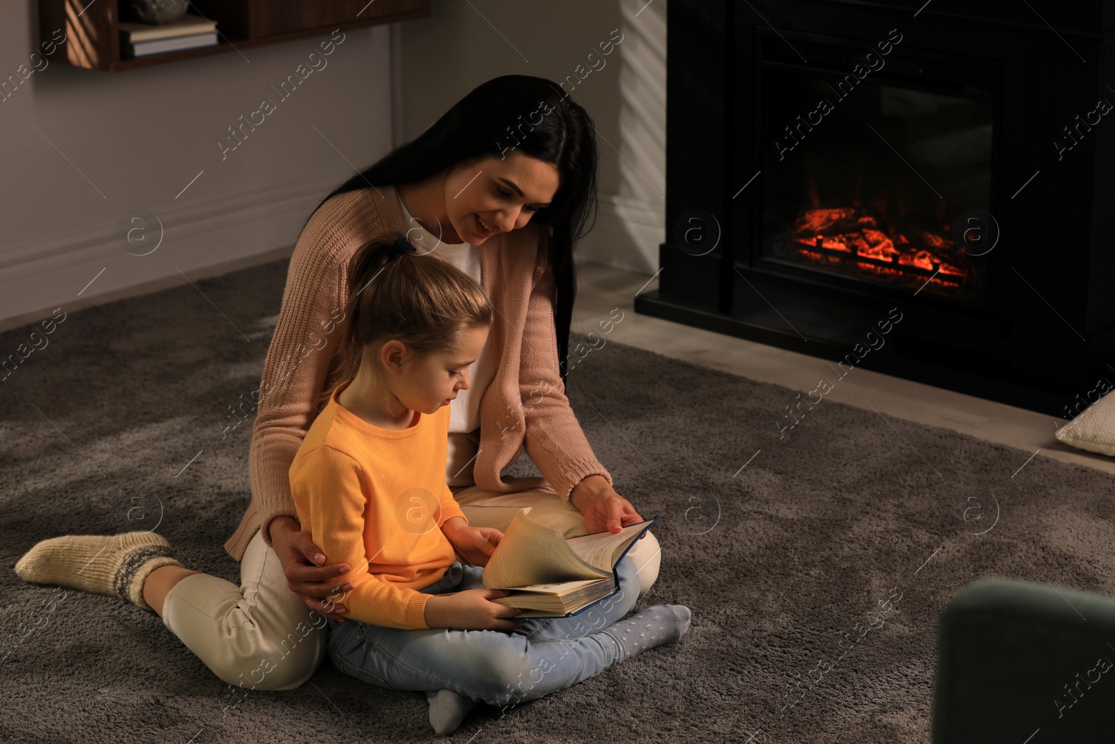 Photo of Happy mother and daughter reading together on floor near fireplace at home