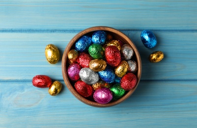 Bowl with chocolate eggs wrapped in colorful foil on light blue wooden table, flat lay