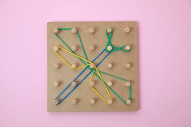 Photo of Wooden geoboard with dragonfly shape made of rubber bands on pink background, top view. Educational toy for motor skills development