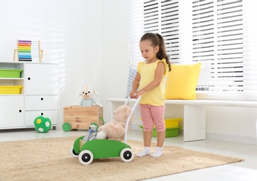 Photo of Cute little girl playing with toy walker and teddy bear at home