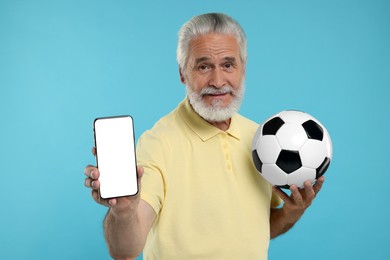 Senior sports fan with soccer ball showing smartphone on light blue background