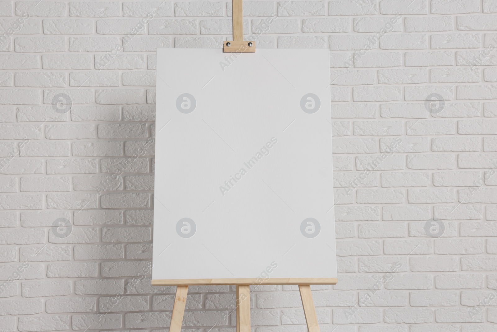 Photo of Wooden easel with blank canvas near white brick wall. Space for text