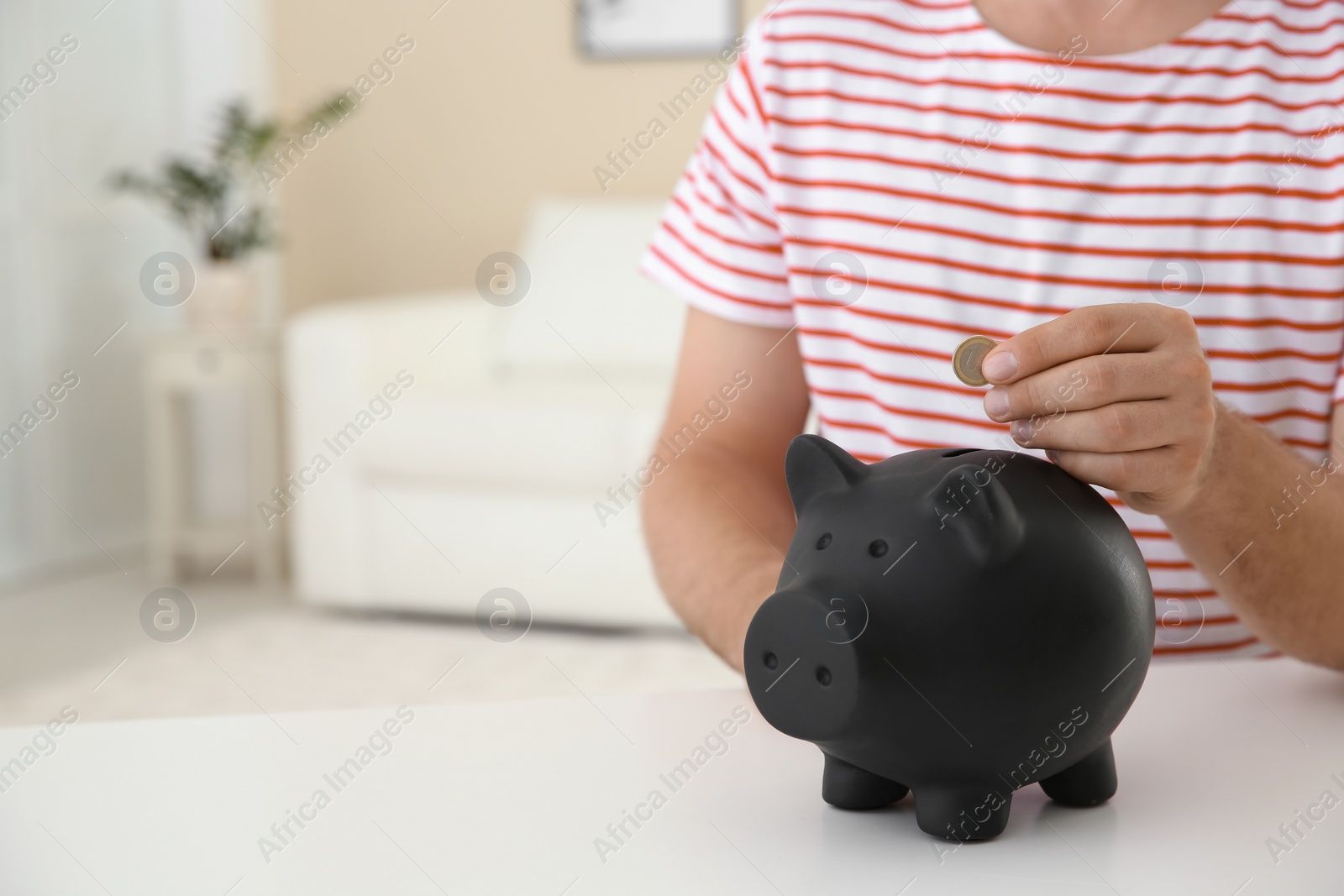 Photo of Man putting coin into piggy bank on table against blurred background, closeup with space for text