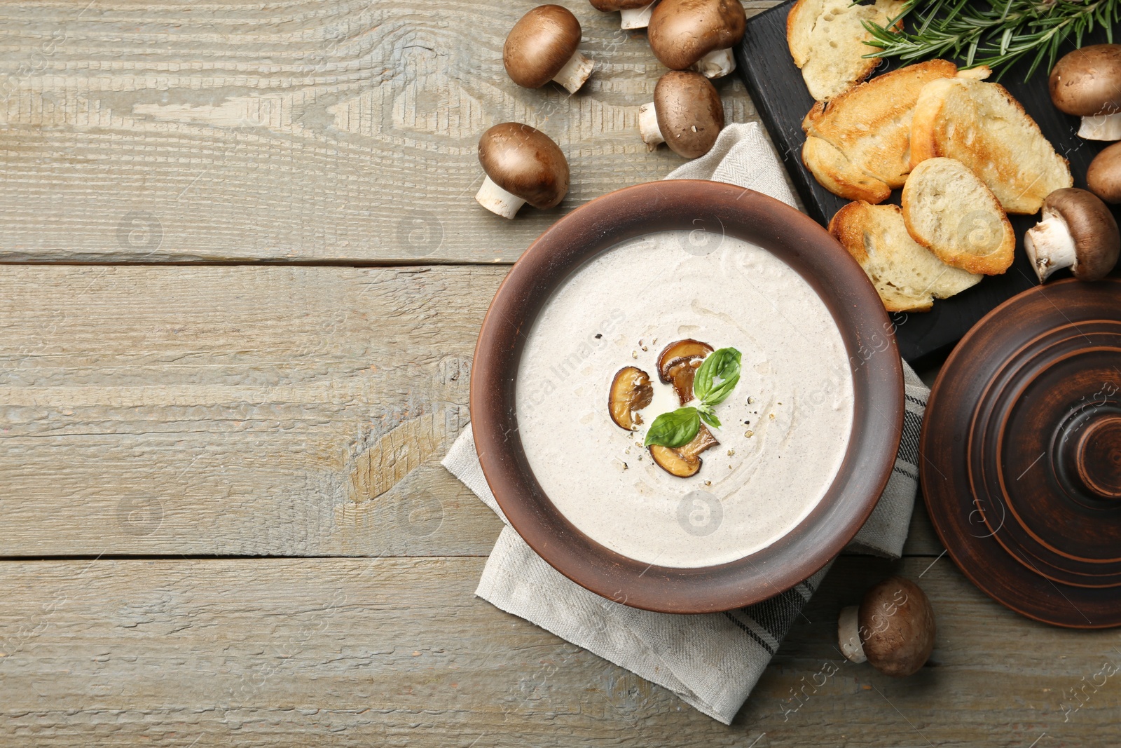 Photo of Tasty homemade mushroom soup in ceramic pot, croutons, fresh champignons and rosemary on wooden table, flat lay. Space for text