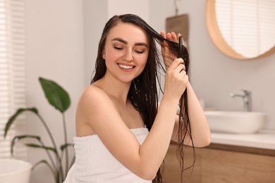 Photo of Young woman brushing hair after applying mask in bathroom