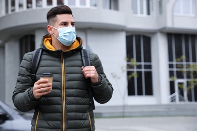 Photo of Man in medical face mask with cup of coffee walking outdoors. Personal protection during COVID-19 pandemic