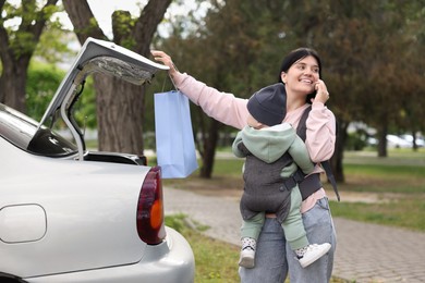 Photo of Mother holding her child in sling (baby carrier) while closing car trunk outdoors