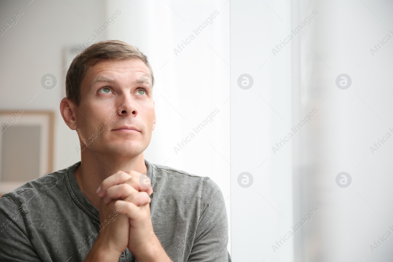 Photo of Man with hands clasped together for prayer near window. Space for text