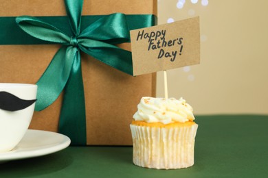 Photo of Happy Father's Day. Tasty cupcake with greetings and gift box on green table, closeup