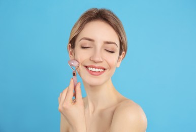 Young woman using natural rose quartz face roller on light blue background