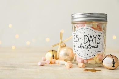 Photo of Glass jar with label 25 Days of Christmas and tasty marshmallows on white wooden table against blurred background, space for text. Advent calendar