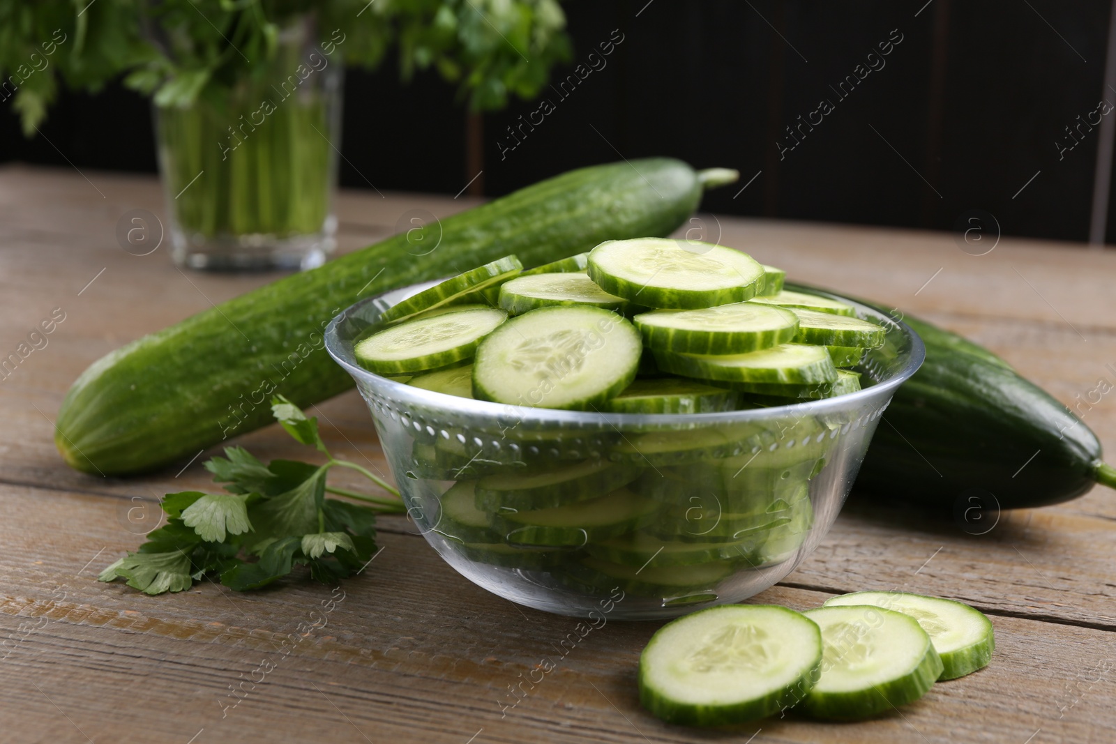 Photo of Cut cucumber in glass bowl, fresh vegetables and parsley on wooden table, closeup