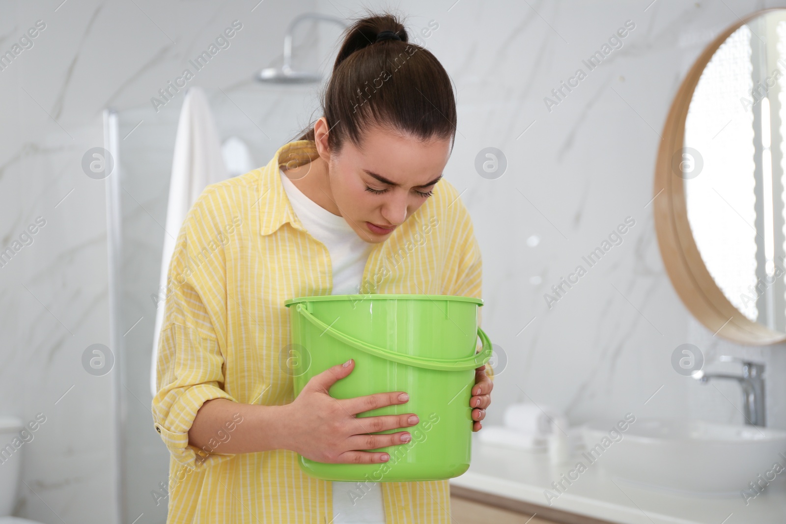 Photo of Young woman with bucket suffering from nausea in bathroom. Food poisoning