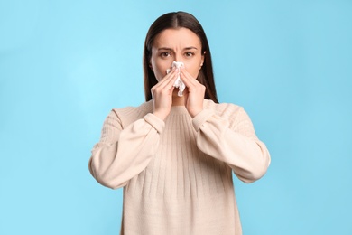 Young woman with tissue suffering from runny nose on light blue background