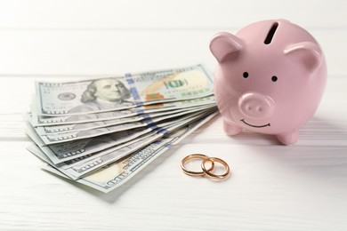 Honeymoon concept. Piggy bank, dollar banknotes and golden rings on white wooden table, closeup