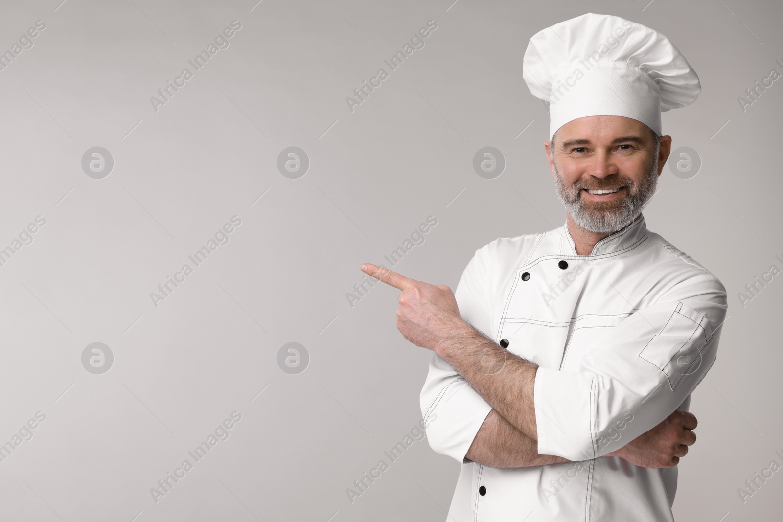 Photo of Happy chef in uniform pointing at something on grey background, space for text