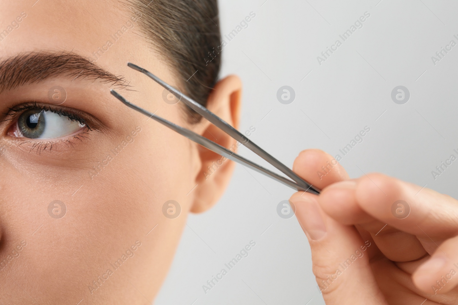 Photo of Eyebrow correction. Young woman with tweezers on light grey background, closeup