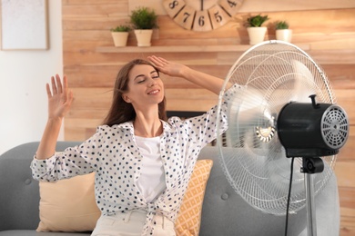 Photo of Woman enjoying air flow from fan on sofa in living room. Summer heat