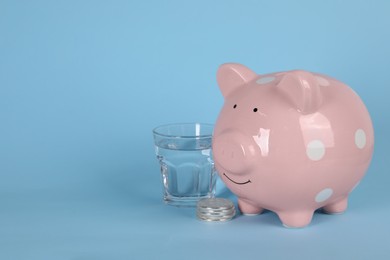 Photo of Water scarcity concept. Piggy bank, coins and glass of drink on light blue background, space for text