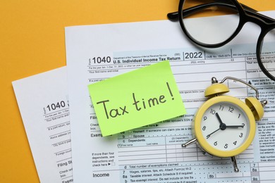 Photo of Reminder note with words Tax Time, documents, glasses and alarm clock on orange background, flat lay