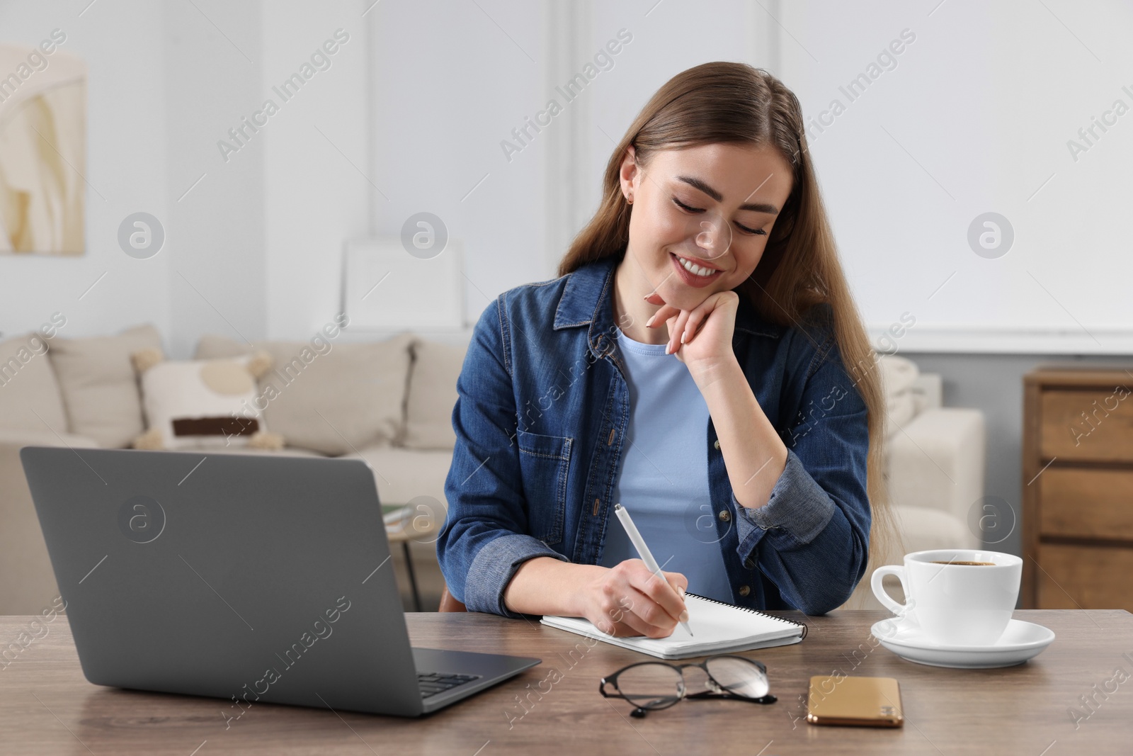 Photo of Happy woman writing something in notebook near laptop at wooden table in room