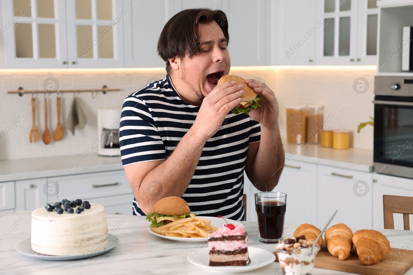 Photo of Hungry overweight man eating tasty burger at table in kitchen