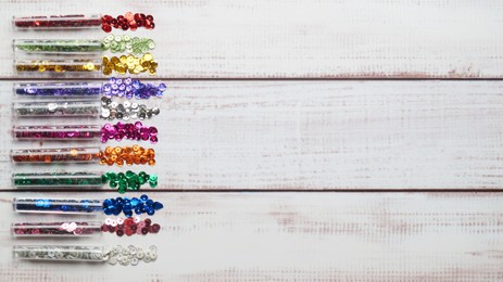 Photo of Tubes with many colorful sequins on white wooden table, flat lay. Space for text