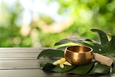 Image of Golden singing bowl, mallet, green leaf and flower on wooden table outdoors, space for text